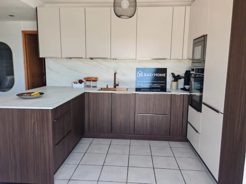 Gros plan sur l'îlot central en bois foncé d'une cuisine moderne, plan de travail effet marbre blanc, évier encastré et mitigeur de couleur cuivre, bol de fruits sur l'îlot, plaque EasyHome et appareils électroménagers en arrière-plan.