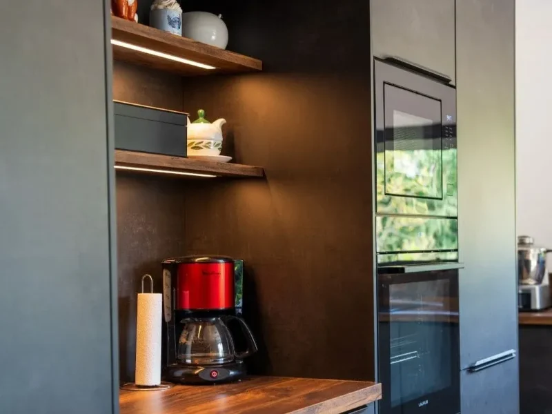 Mur de colonnes de cuisine noires mat avec électroménager encastré (micro-ondes, four), niches lumineuses avec étagères en bois exposant une machine à café rouge, de la décoration et un dévidoir à essuie-tout.