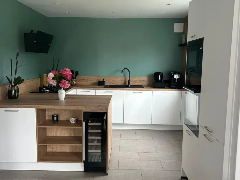 Vue perspective centrale d'une cuisine scandinave avec des meubles blancs mats, un plan de travail et une crédence en bois clair, un mur peint en vert sauge, un évier noir avec mitigeur noir devant, un îlot central avec étagères bois et une cave à vin encastrée, un vase de fleurs roses sur l'îlot.
